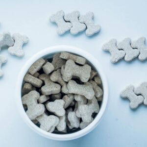 Top view of bone-shaped dog treats in a white bowl, on a light blue background.